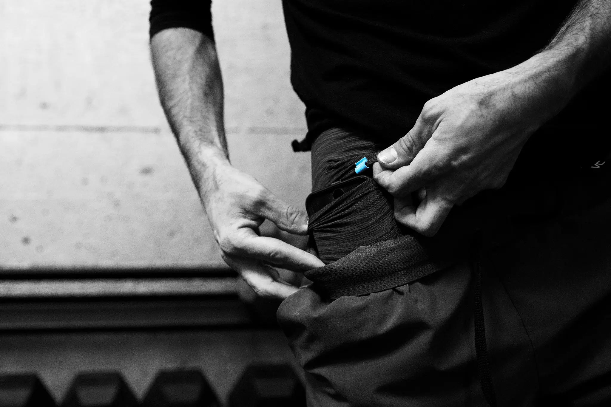 Close-up black and white action shot of a person demonstrating the secure fold pocket on Pacterra Men’s Middy Compression Shorts, showing a phone being tucked into the side compartment during a workout.