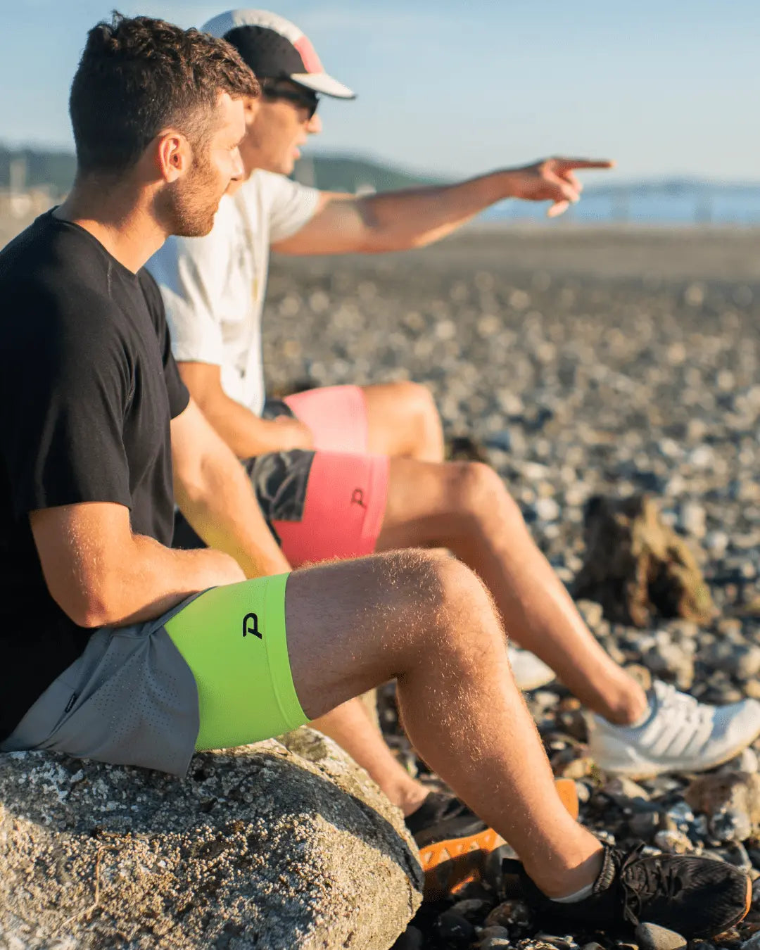 Two athletes sitting on a rocky beach wearing Pacterra Middy compression shorts in volt yellow and pink, demonstrating the comfortable, non-restrictive fit during rest.