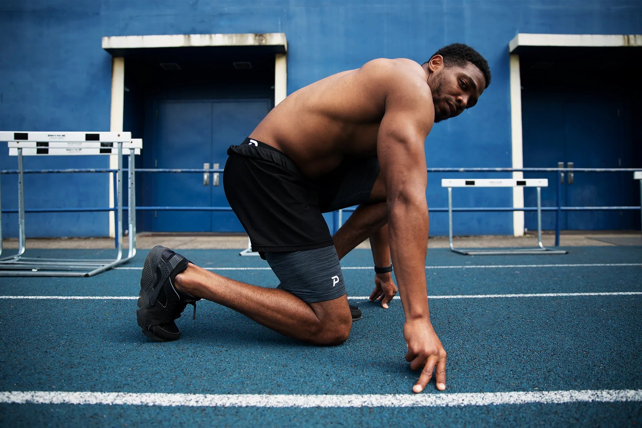Athlete in a sprinter's starting stance on a blue track wearing charcoal grey Pacterra Middy compression shorts under black training shorts.