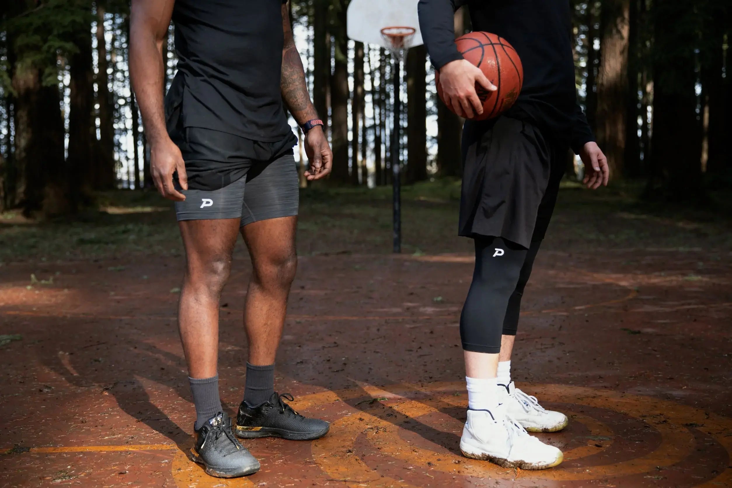 Two athletes on an outdoor basketball court in the woods; one wearing grey Middy compression shorts and the other wearing black Strider tights.