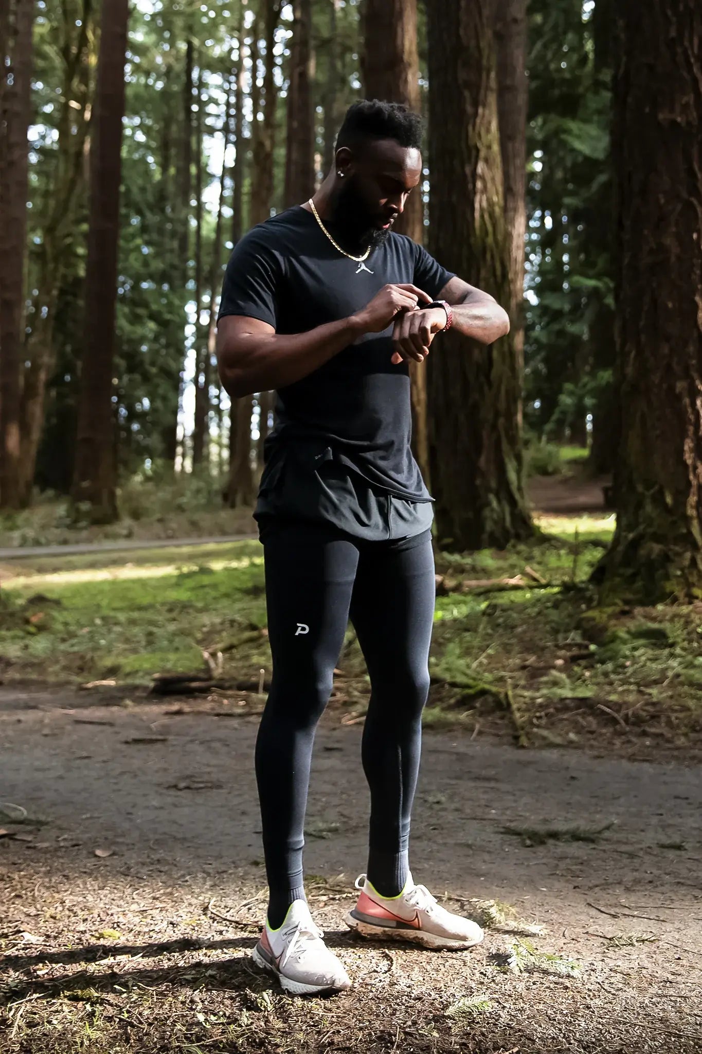 Athlete standing on a wooded trail wearing black Pacterra Strider compression tights, checking his smartwatch during an outdoor training session.