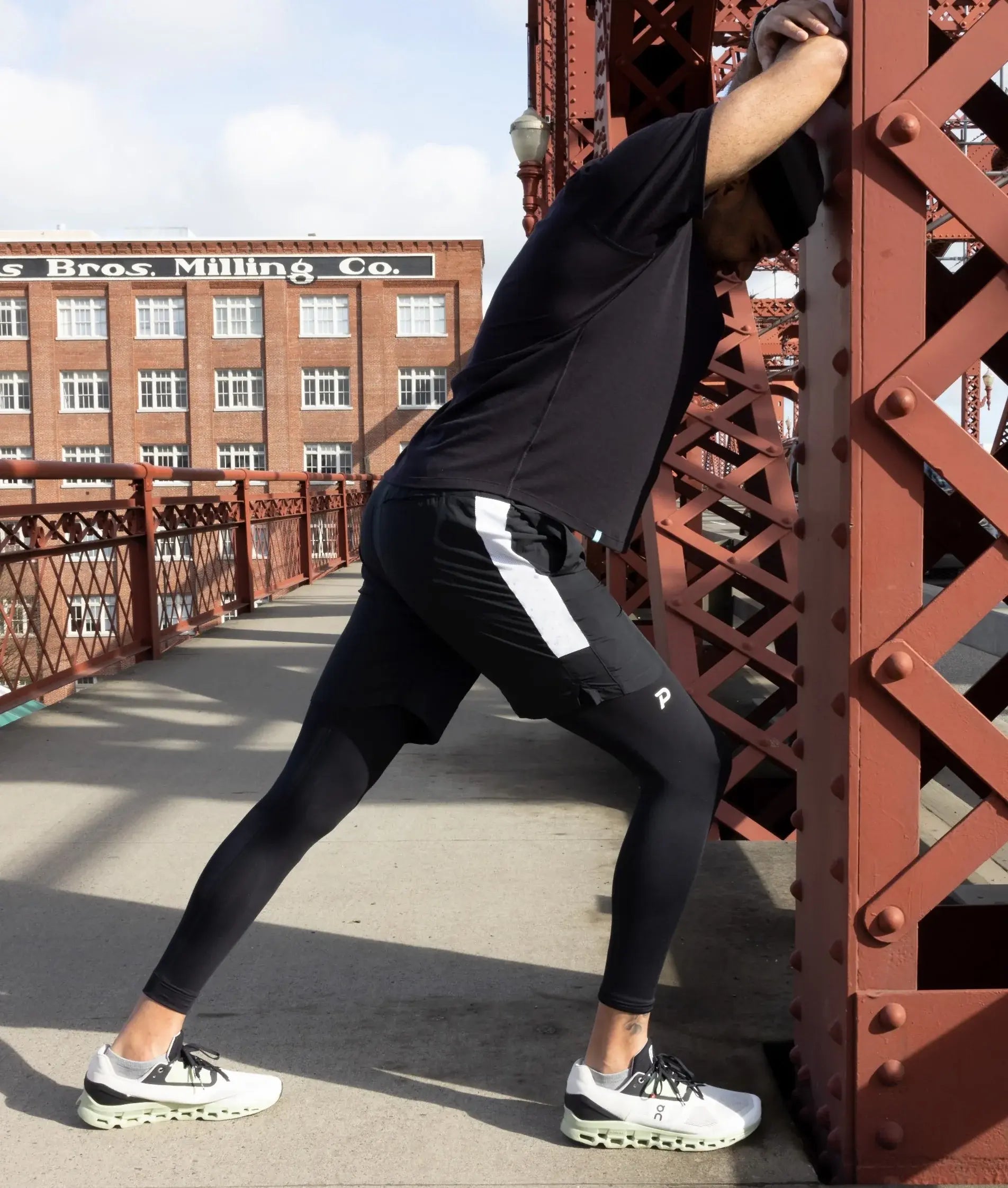 Athlete wearing black Pacterra Strider compression tights stretching against a red bridge structure in an urban setting with a historic brick building background.