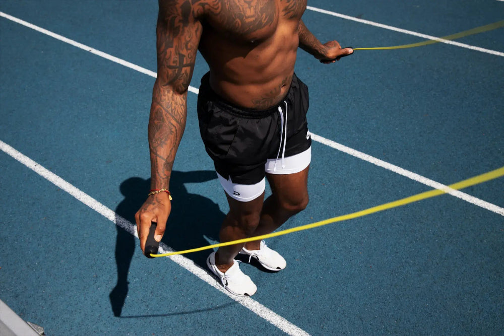 Male athlete jumping rope on a blue track wearing Pacterra Middy performance compression shorts in white under black training shorts. The image shows the secure fit and high-mobility design of Pacterra athletic wear.