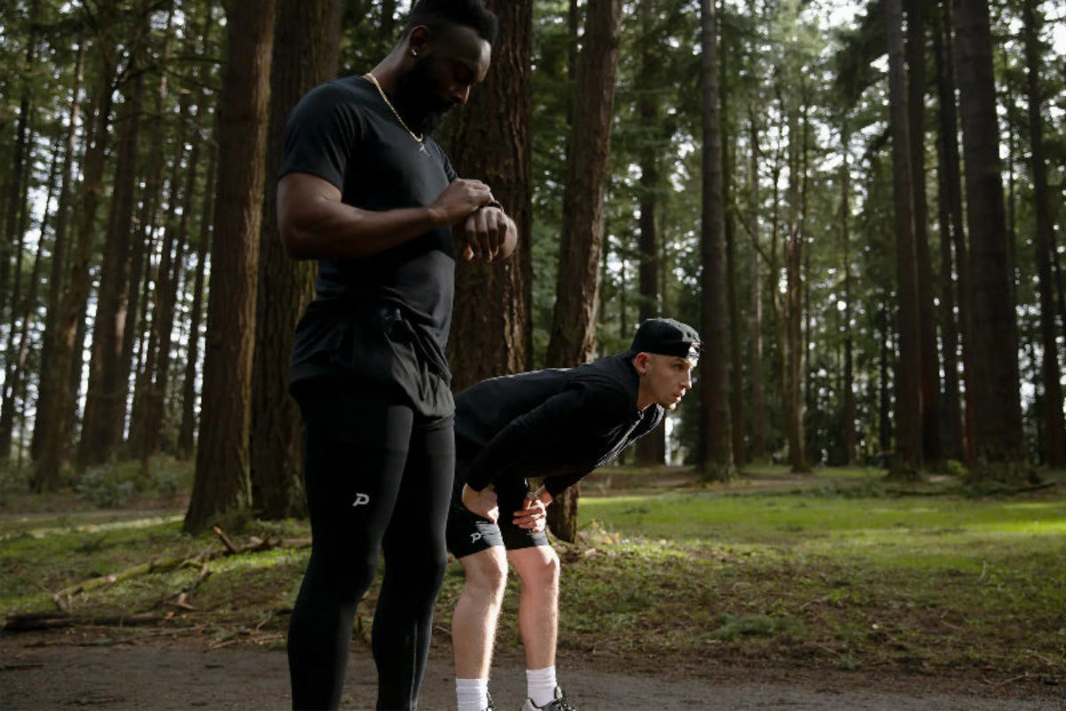 Two athletes wearing Pacterra compression leggings and shorts during a trail run in the forest, tracking performance on a smartwatch.