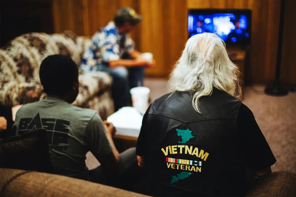 A Vietnam Veteran wearing a black leather vest with service ribbons, sitting with other veterans during a Pacterra Athletics donation event.