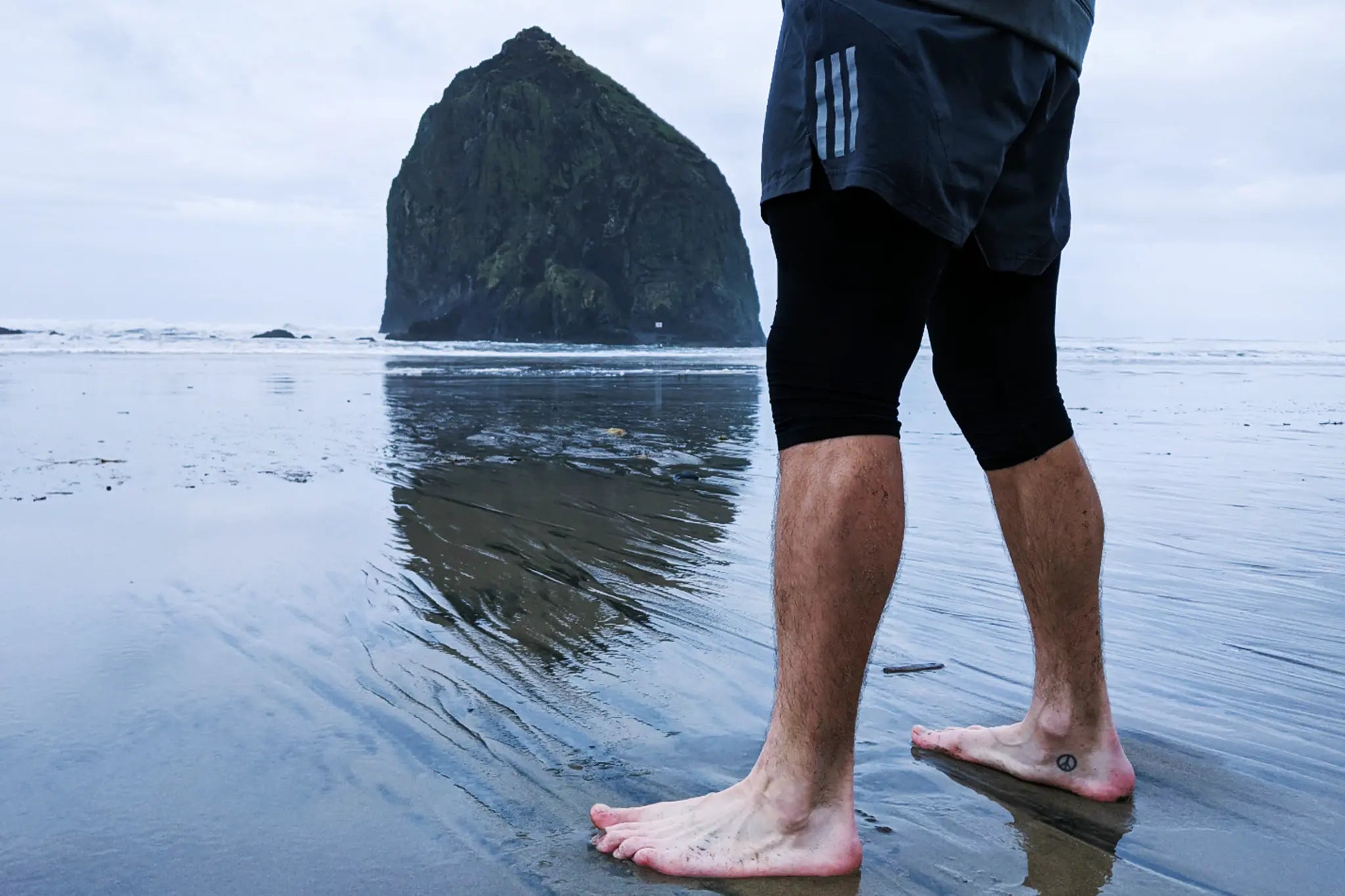 A man standing barefoot on Cannon Beach in front of Haystack Rock wearing Pacterra mens 3/4 Strider compression tights.