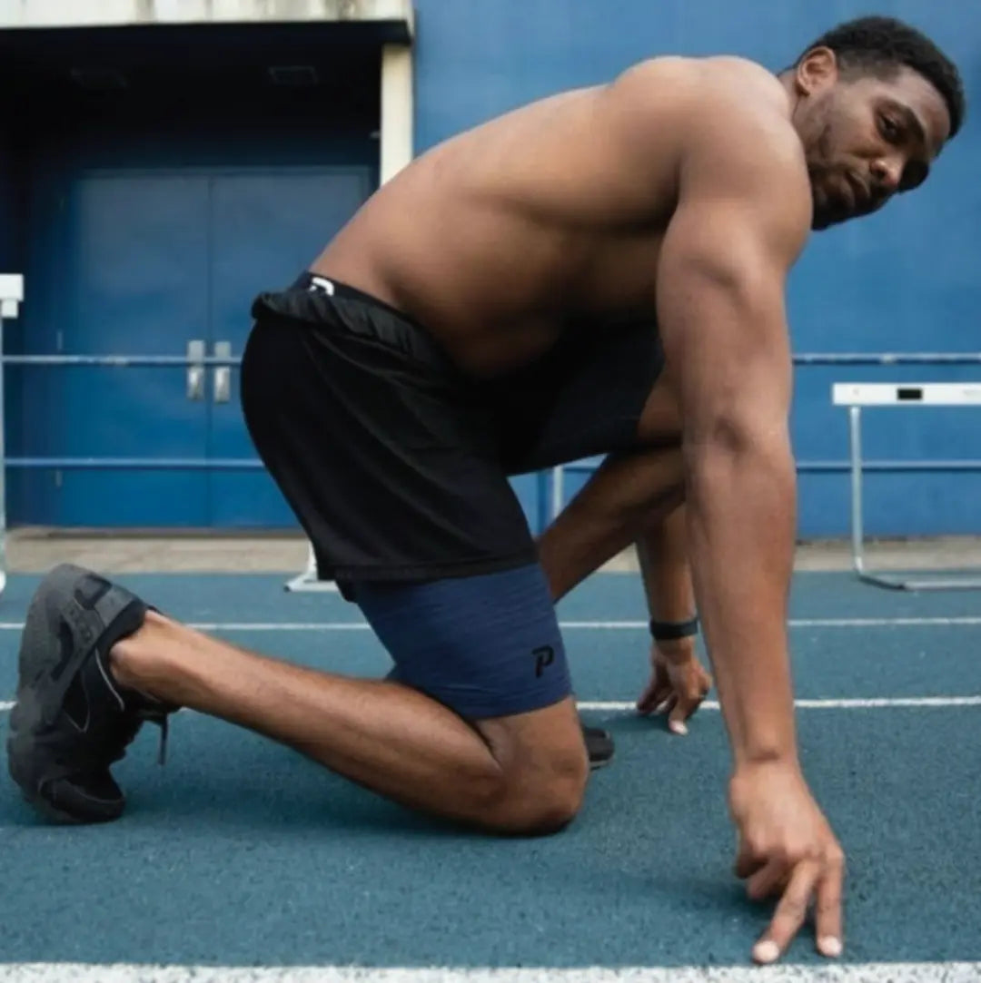 An athlete in a sprinter's start position on a track wearing navy Middy compression shorts under gym shorts, illustrating the secure fit and muscle support for explosive movements.