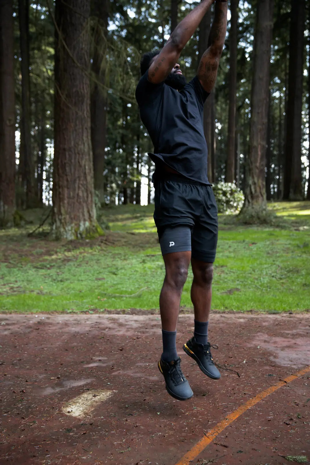 Athlete performing a vertical jump in a wooded outdoor setting wearing heather grey Pacterra Middy compression shorts, showing the stay-put fit during explosive plyometric training.
