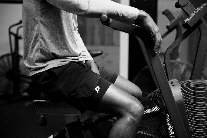 Black and white action shot of an athlete wearing black Pacterra Middy compression shorts while training on an air bike, highlighting the non-slip performance fit.